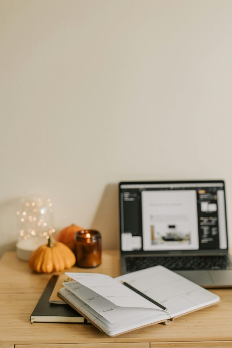 Warm and inviting workspace with open planner, laptop, candle, and pumpkins for a fall decorative touch.
